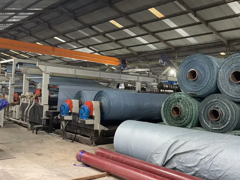 Mesin laminasi terpal dengan tumpukan roll hijau dan biru di pabrik YO TERPAL NUSANTARA RAYA